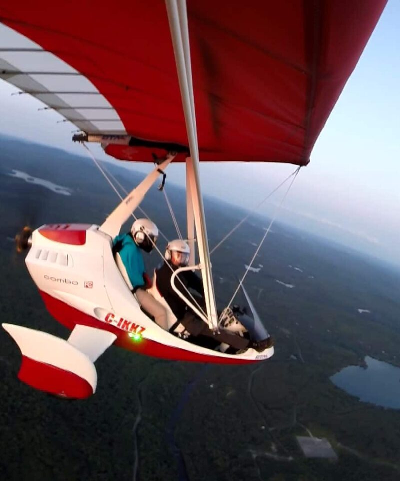 A person is flying a red and white ultralight aircraft with a passenger over a landscape of forests and lakes at sunset.
