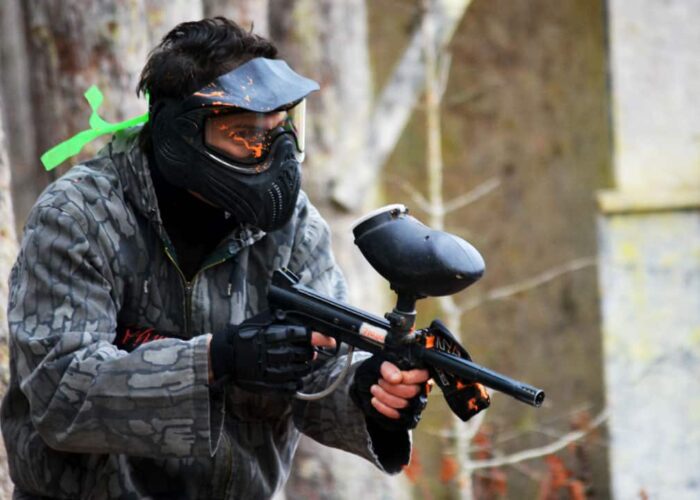 Person wearing camouflage and a protective mask holds a paintball gun, standing outdoors near trees and a building.