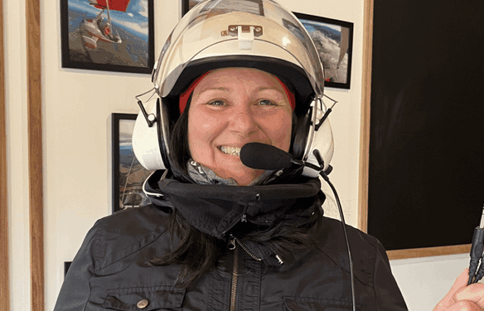 A person wearing a pilot helmet with a microphone stands indoors, smiling at the camera. Framed aviation photos are visible on the wall behind them.
