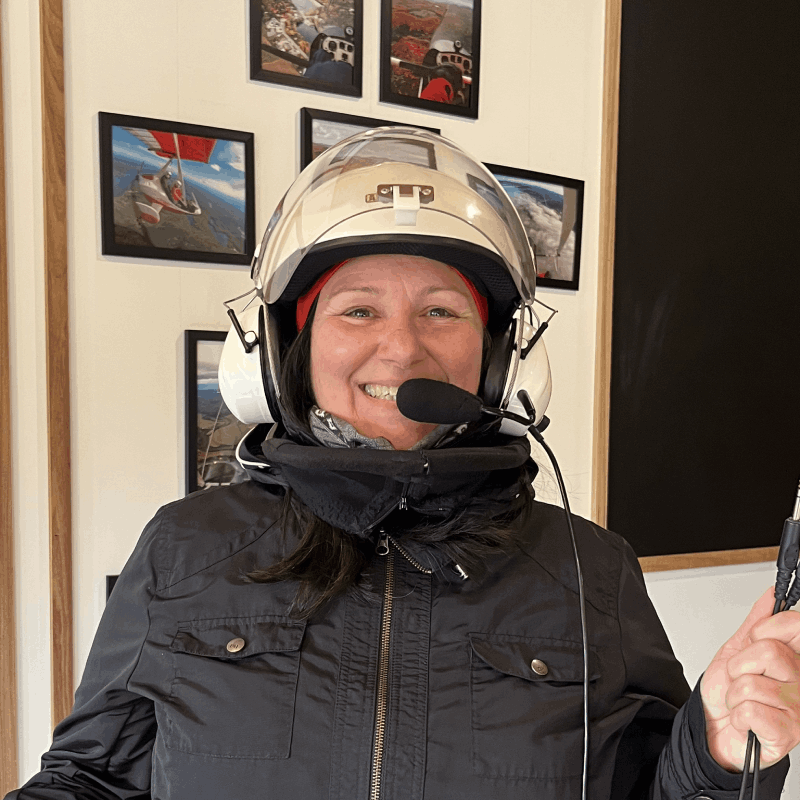 A person wearing a pilot helmet with a microphone stands indoors, smiling at the camera. Framed aviation photos are visible on the wall behind them. A person wearing a pilot helmet with a microphone stands indoors, smiling at the camera. Framed aviation photos are visible on the wall behind them.
