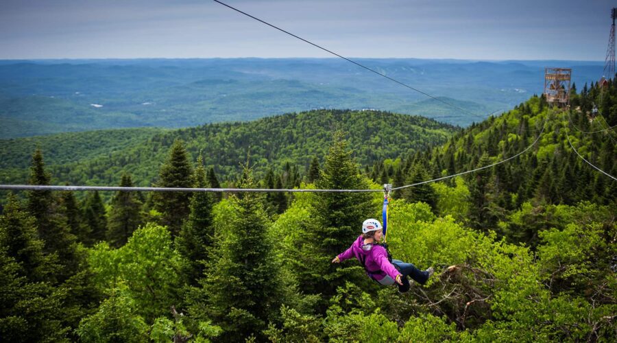 A person wearing a helmet and harness rides a zipline above a dense green forest with hills and a cloudy sky in the background. A person wearing a helmet and harness rides a zipline above a dense green forest with hills and a cloudy sky in the background.