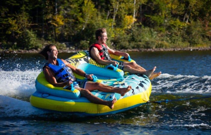 Deux hommes portant des gilets de sauvetage font une bouée tractée sur un lac, tirés par un bateau, avec des arbres en arrière-plan - une façon amusante de découvrir les sports nautiques.