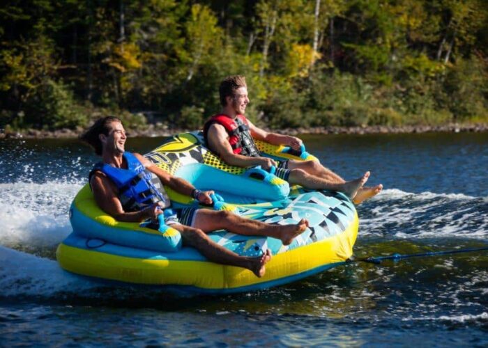 Two men wearing life jackets ride a large inflatable towable tube on a lake, being pulled by a boat, with trees visible in the background.