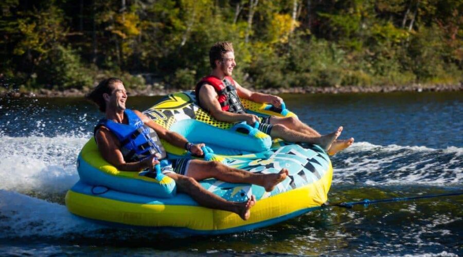 Deux hommes portant des gilets de sauvetage font une bouée tractée sur un lac, tirés par un bateau, avec des arbres en arrière-plan – une façon amusante de découvrir les sports nautiques. Deux hommes portant des gilets de sauvetage font une bouée tractée sur un lac, tirés par un bateau, avec des arbres en arrière-plan - une façon amusante de découvrir les sports nautiques.