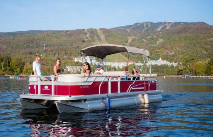 A group of people sit and stand on a red pontoon boat on a calm lake, with tree-covered hills and clear skies in the background.