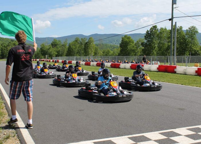 A person waves a green flag as a group of go-kart drivers, all wearing helmets, start a race on an outdoor track surrounded by trees and mountains.