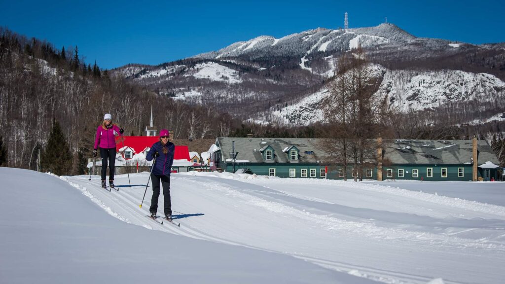 Two people cross-country skiing on a snowy trail with snow-covered mountains and buildings in the background under a clear blue sky.
