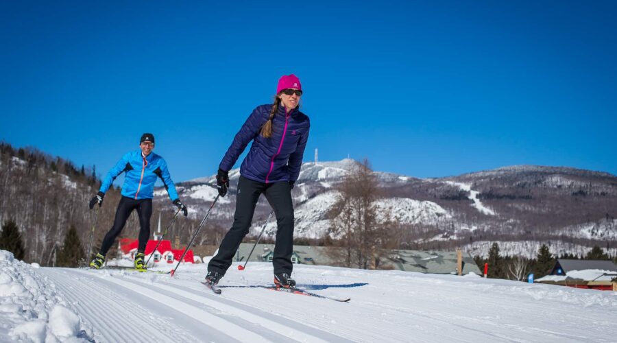Two people cross-country skiing on a groomed snowy trail with mountains and blue sky in the background. Two people cross-country skiing on a groomed snowy trail with mountains and blue sky in the background.