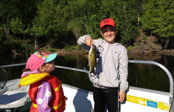 A boy in a red cap holds up a fish on a boat while a younger girl in a pink cap and life jacket looks at him. Trees and water are visible in the background.
