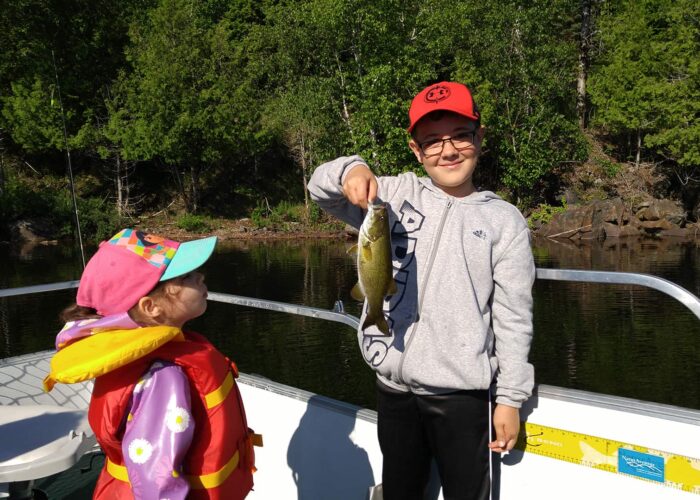 A boy in a red cap holds up a fish on a boat while a younger girl in a pink cap and life jacket looks at him. Trees and water are visible in the background.