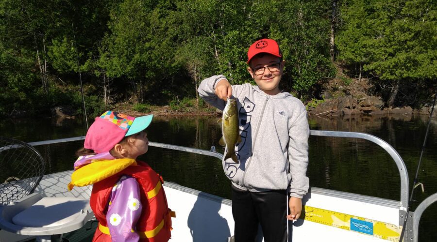 Deux enfants sur un bateau au lac Ouimet : l’un brandit un poisson et sourit à l’appareil photo, tandis que l’autre, vêtu d’un gilet de sauvetage, regarde le poisson. Les arbres et l’eau servent de décor à ce moment amusant de pêche en famille. Deux enfants sur un bateau au lac Ouimet : l'un brandit un poisson et sourit à l'appareil photo, tandis que l'autre, vêtu d'un gilet de sauvetage, regarde le poisson. Les arbres et l'eau servent de décor à ce moment amusant de pêche en famille.