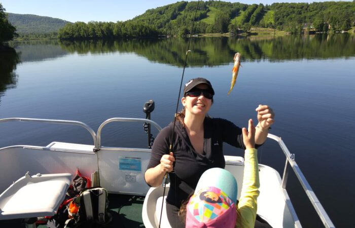 A woman on a boat holds a fishing rod with a small fish on the line while a child in a colorful hat reaches toward it; a calm lake and green hills are in the background.