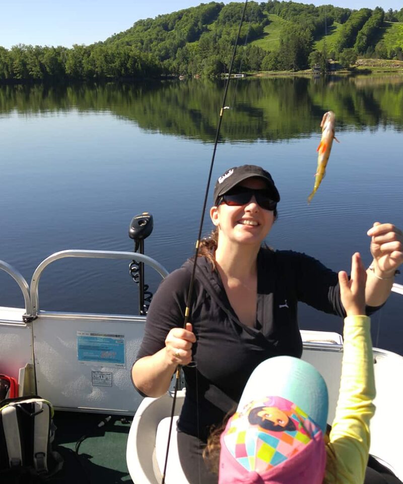 A woman on a boat holds a fishing rod with a small fish on the line while a child in a colorful hat reaches toward it; a calm lake and green hills are in the background.