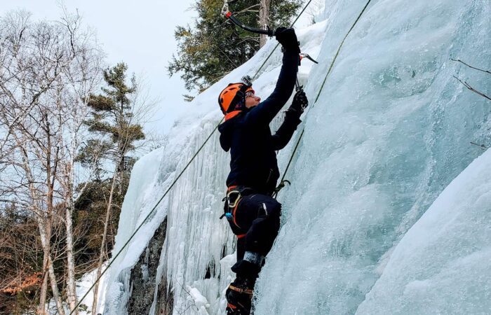 Person wearing winter gear and helmet climbs a vertical ice wall using ice axes and safety ropes; snow-covered trees are visible in the background.