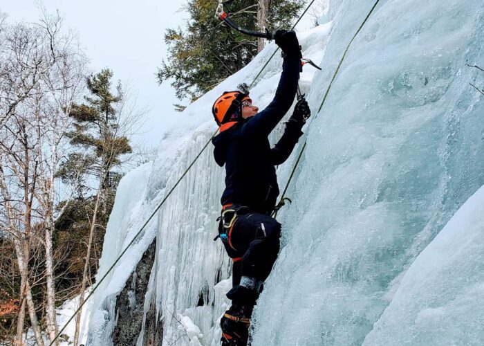 Person wearing winter gear and helmet climbs a vertical ice wall using ice axes and safety ropes; snow-covered trees are visible in the background.