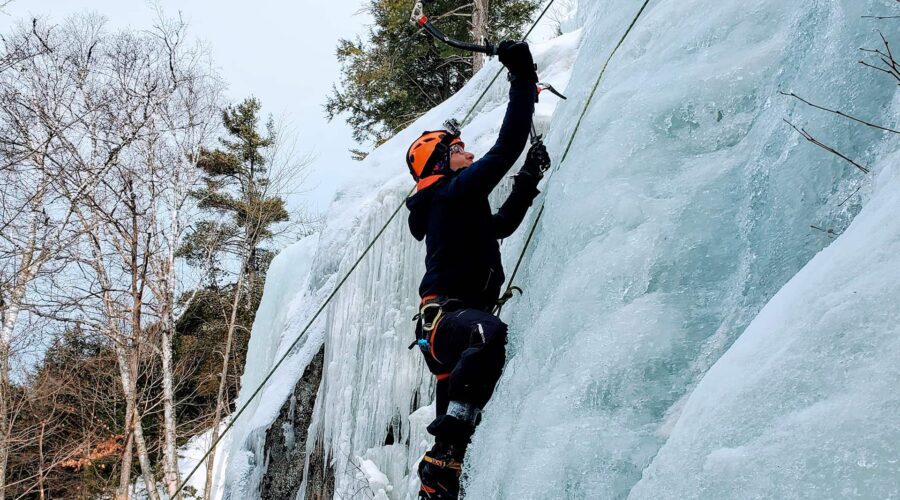 Person wearing winter gear and helmet climbs a vertical ice wall using ice axes and safety ropes; snow-covered trees are visible in the background. Person wearing winter gear and helmet climbs a vertical ice wall using ice axes and safety ropes; snow-covered trees are visible in the background.
