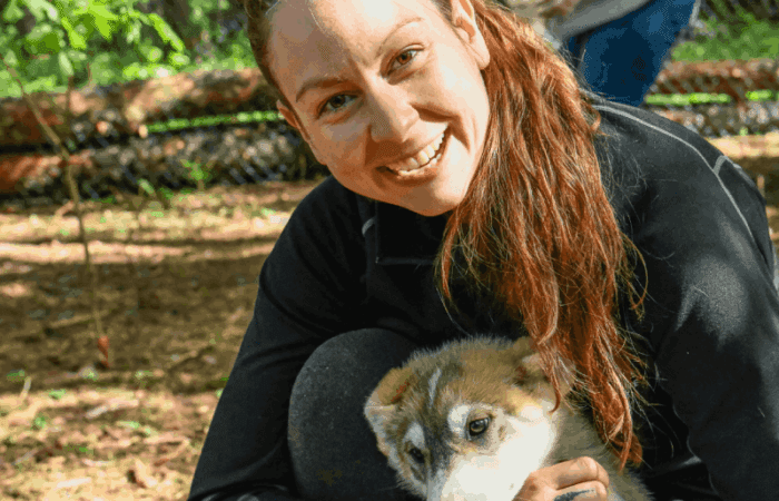 A woman crouches outdoors, smiling and holding a young, light brown and white puppy. Trees and another person appear in the blurred background.