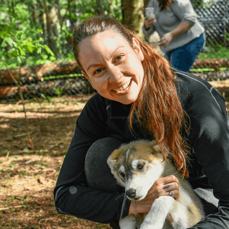 A woman crouches outdoors, smiling and holding a young, light brown and white puppy. Trees and another person appear in the blurred background. A woman crouches outdoors, smiling and holding a young, light brown and white puppy. Trees and another person appear in the blurred background.