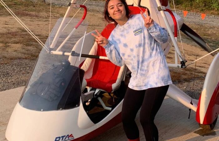 Person standing in front of a small red and white ultralight aircraft, smiling and making peace signs with both hands.