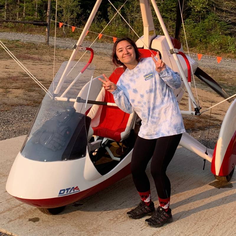 Person standing in front of a small red and white ultralight aircraft, smiling and making peace signs with both hands. Person standing in front of a small red and white ultralight aircraft, smiling and making peace signs with both hands.