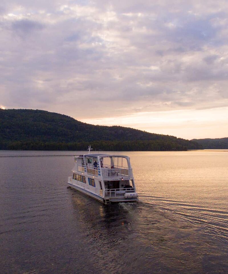 A white boat moves across a calm lake with forested hills and a cloudy sky in the background during sunset.