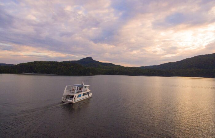 A white tour boat travels across a calm lake with forested hills and a cloudy sky in the background.