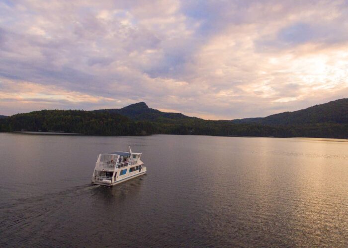 A white tour boat travels across a calm lake with forested hills and a cloudy sky in the background.