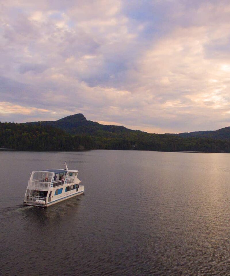 Un bateau blanc d'un service de location de bateaux navigue sur un lac calme, entouré de collines boisées, sous un ciel nuageux au coucher du soleil.
