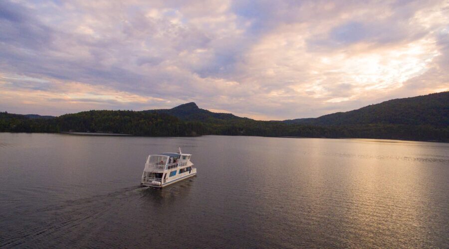 Un bateau blanc d’un service de location de bateaux navigue sur un lac calme, entouré de collines boisées, sous un ciel nuageux au coucher du soleil. Un bateau blanc d'un service de location de bateaux navigue sur un lac calme, entouré de collines boisées, sous un ciel nuageux au coucher du soleil.