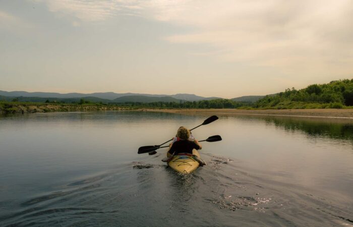 Person paddling a yellow kayak on a calm river surrounded by trees and distant mountains under a cloudy sky.