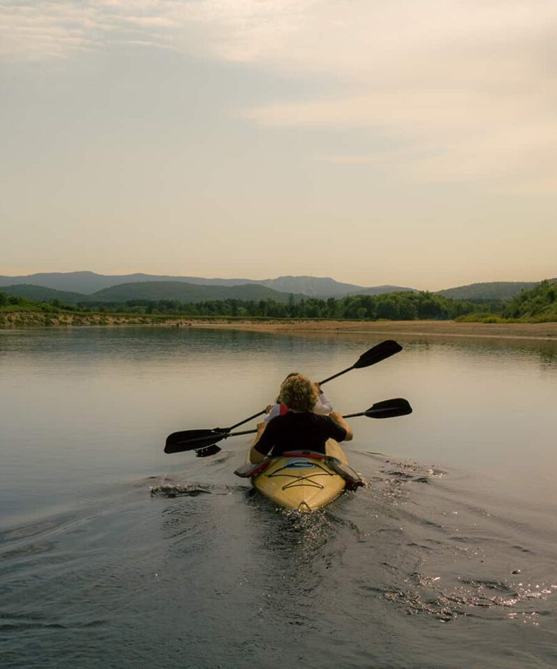 Person paddling a yellow kayak on a calm river surrounded by trees and distant mountains under a cloudy sky.