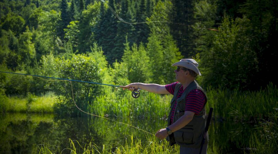 A person wearing a hat and fishing vest is fly fishing by a pond surrounded by green trees and tall grass. A person wearing a hat and fishing vest is fly fishing by a pond surrounded by green trees and tall grass.