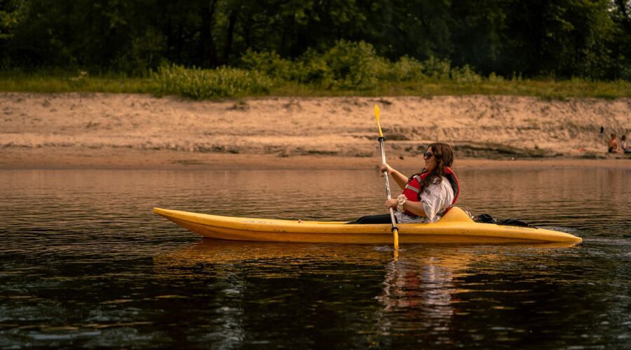A person wearing a life jacket paddles a yellow kayak on calm water near a sandy shore with trees in the background. A person wearing a life jacket paddles a yellow kayak on calm water near a sandy shore with trees in the background.