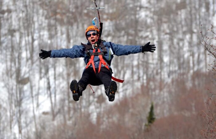 Person wearing winter gear and helmet ziplining outdoors with snowy, leafless trees in the background, arms outstretched.