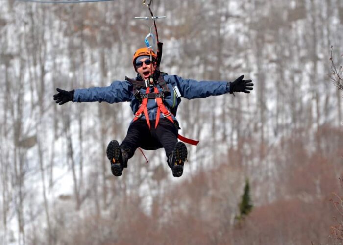 Person wearing winter gear and helmet ziplining outdoors with snowy, leafless trees in the background, arms outstretched.