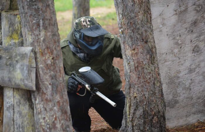 A person wearing paintball gear and a mask crouches behind a tree, holding a paintball marker during an outdoor game.