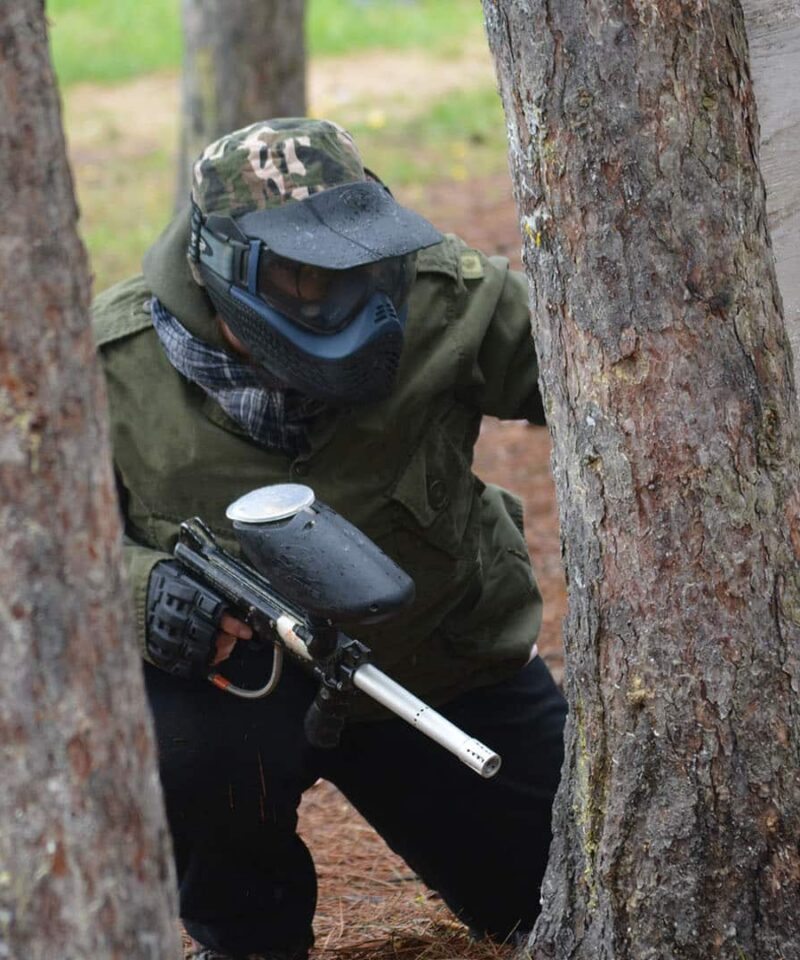 A person wearing paintball gear and a mask crouches behind a tree, holding a paintball marker during an outdoor game.