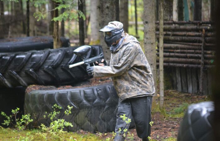 A person wearing camouflage and protective gear holds a paintball gun while standing near large tires in a wooded outdoor area.