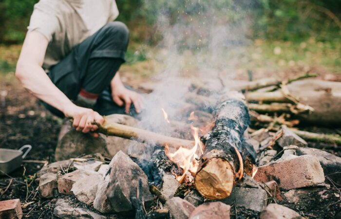 A person crouches by a campfire made of logs and surrounded by rocks and bricks, tending the fire with a stick in an outdoor wooded area.