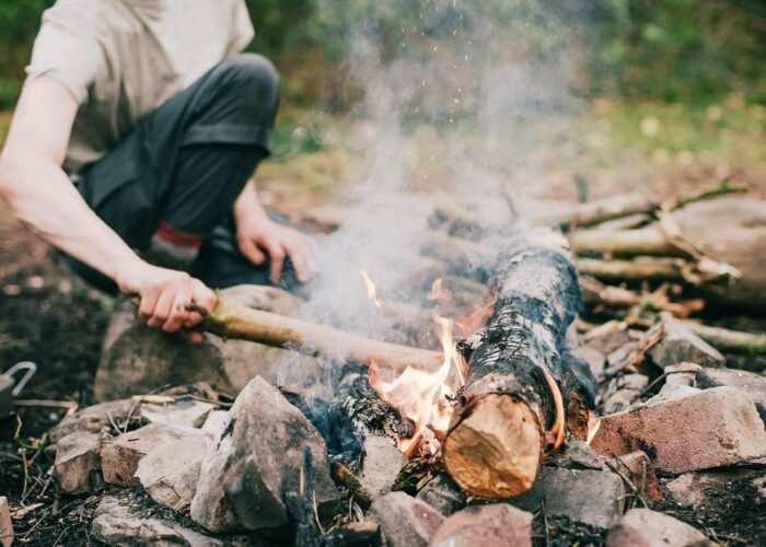 A person crouches by a campfire made of logs and surrounded by rocks and bricks, tending the fire with a stick in an outdoor wooded area.