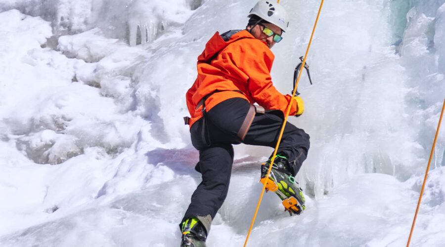 A person wearing an orange jacket, helmet, and climbing gear ascends an icy slope using crampons and an ice axe, secured by a rope. A person wearing an orange jacket, helmet, and climbing gear ascends an icy slope using crampons and an ice axe, secured by a rope.