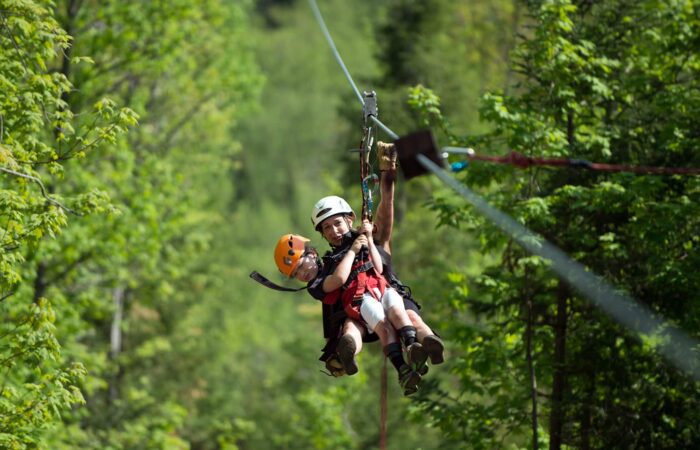 Two people wearing helmets and harnesses ride a zipline through a green forest on a sunny day. Two people wearing helmets and harnesses ride a zipline through a green forest on a sunny day.