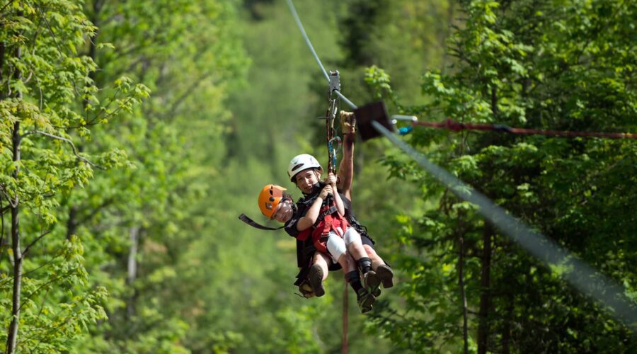 Two people wearing helmets and harnesses ride a zipline through a green forest on a sunny day. Two people wearing helmets and harnesses ride a zipline through a green forest on a sunny day.