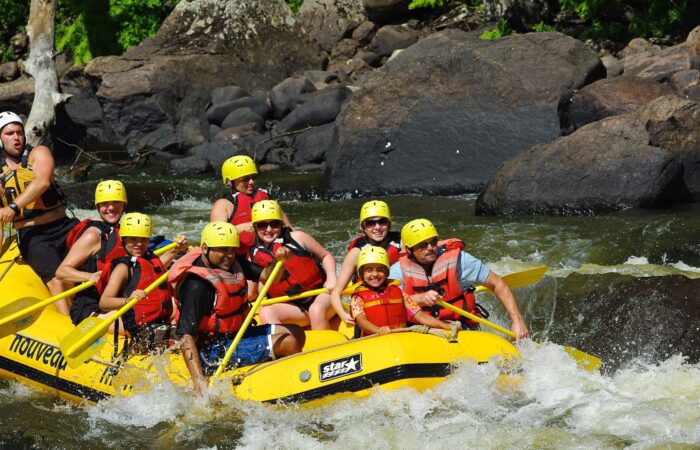 A group of people wearing helmets and life jackets paddle a yellow inflatable raft through whitewater rapids surrounded by rocks.