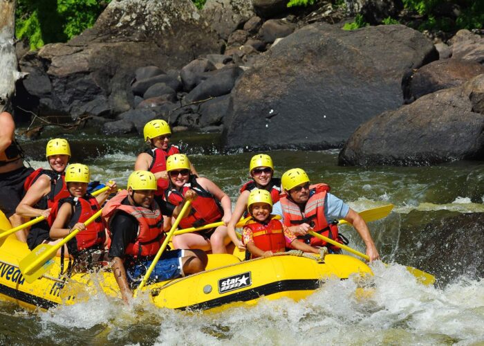 A group of people wearing helmets and life jackets paddle a yellow inflatable raft through whitewater rapids surrounded by rocks.