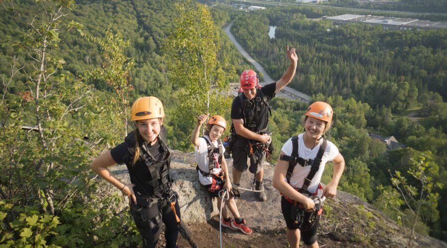 Four people wearing helmets and harnesses stand on a rocky cliff edge overlooking a forested valley and industrial buildings in the distance. Four people wearing helmets and harnesses stand on a rocky cliff edge overlooking a forested valley and industrial buildings in the distance.