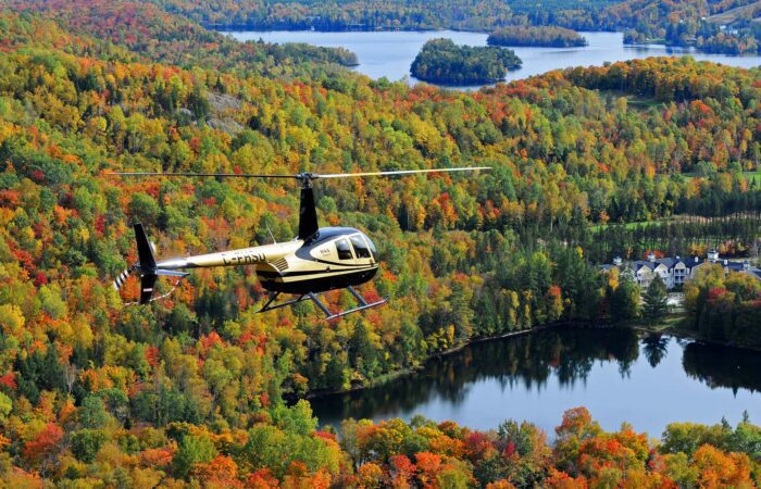 Un hélicoptère de Mont-Tremblant survole un paysage forestier parsemé de lacs, avec des arbres au feuillage automnal dans les tons vert, jaune, orange et rouge.