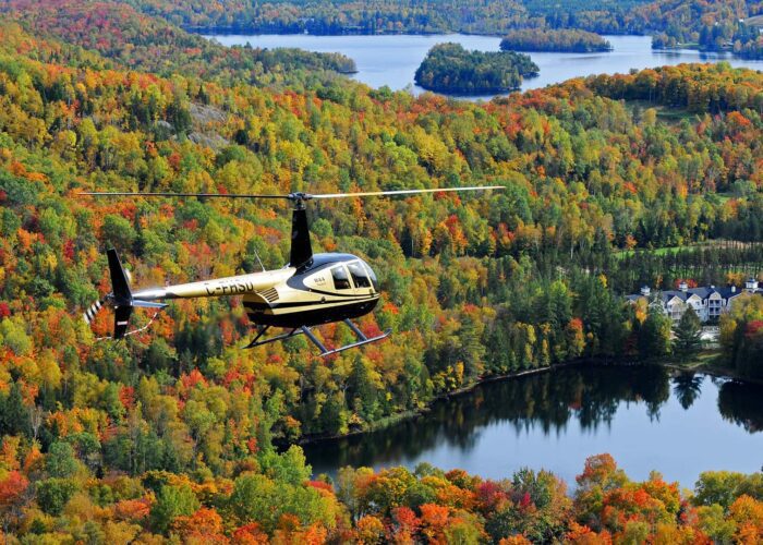 A helicopter flies over a forested landscape with fall foliage in various colors, lakes, and a few buildings visible below.