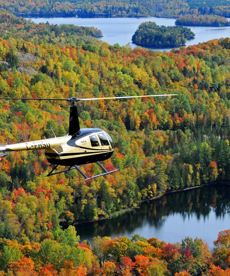 A helicopter flies over a forested landscape with fall foliage in various colors, lakes, and a few buildings visible below.