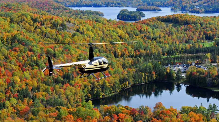 Un hélicoptère de Mont-Tremblant survole un paysage forestier parsemé de lacs, avec des arbres au feuillage automnal dans les tons vert, jaune, orange et rouge.